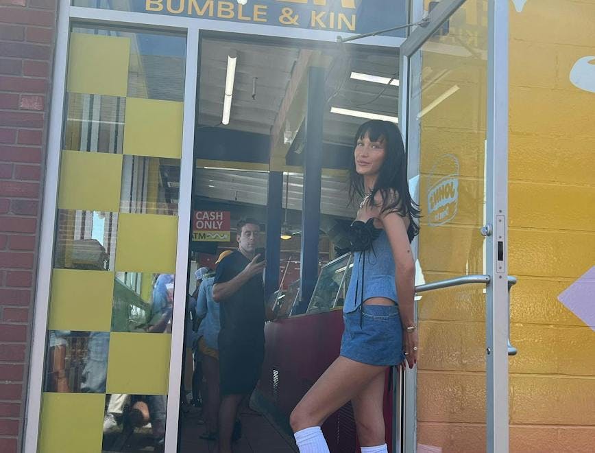 Bella Hadid standing in the doorway of a restaurant called Bella's Diner. She is wearing a denim corset and mini skirt with knee high socks and loafers.